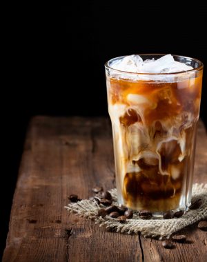 Ice coffee in a tall glass with cream poured over and coffee beans on a old rustic wooden table. Cold summer drink on a dark wooden background with copy space.